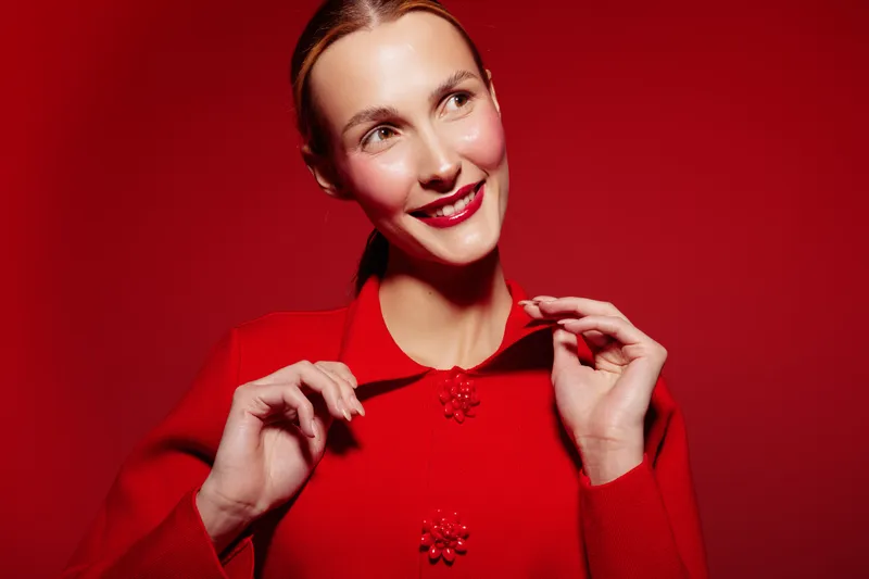 Smiling woman in red sweater poses against red background, looking away with bright lipstick. Concept of fashion, beauty, and vibrant color contrast in studio lighting.
