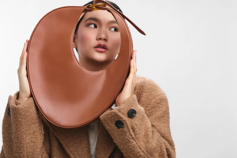 Asian woman wearing a warm coat holds a circular brown accessory to her head, posing in a high fashion studio shot with soft lighting and a thoughtful expression.
