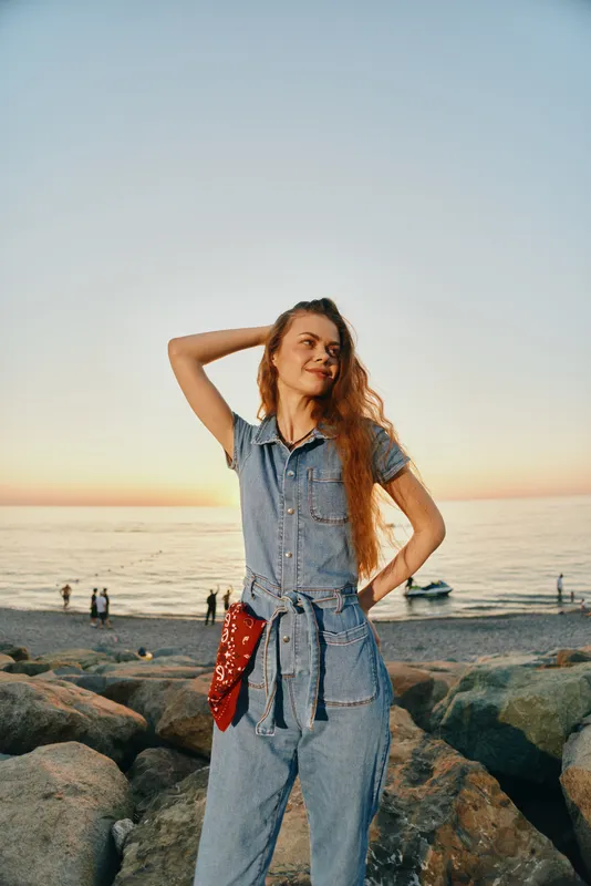 Lifestyle woman in modern boho-western denim street style stands confidently by rocky beach at sunset, showcasing relaxed film color tones and casual summer vibes.
