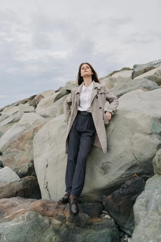 Young woman in stylish coat and black pants stands confidently on rocky shore under cloudy sky. Concept of fashion, confidence, outdoor lifestyle, and autumn style.