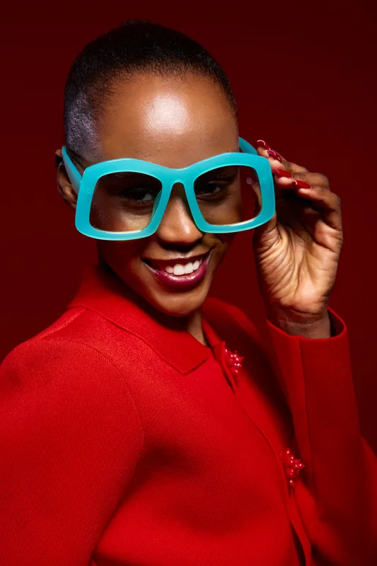 Stylish smiling black African woman with dark skin wearing large turquoise glasses and red jacket on red background. Concept of fashion, confidence, and modern style.