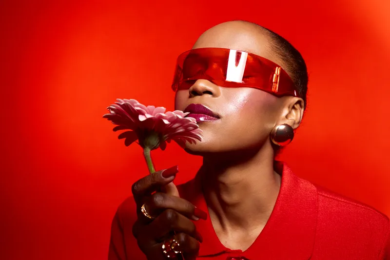 Stylish black woman with dark skin wearing red sunglasses and red outfit holds pink flower near face on red background. Concept for fashion, beauty, and modern portrait photography.