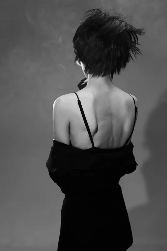 Black and white portrait of a slender woman with short hair showing her back, wearing a dress and jacket, expressing mystery and elegance.