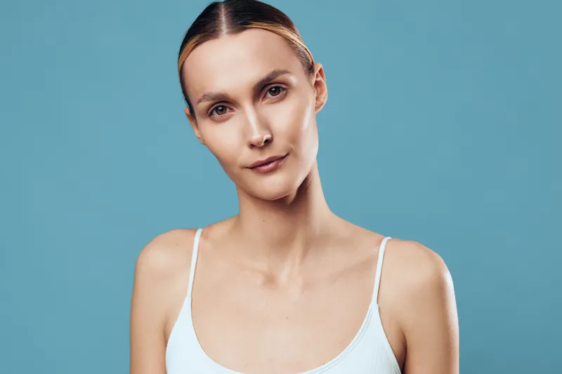 Young woman with long brown hair looks confidently into the camera wearing a white tank top against a plain blue background.