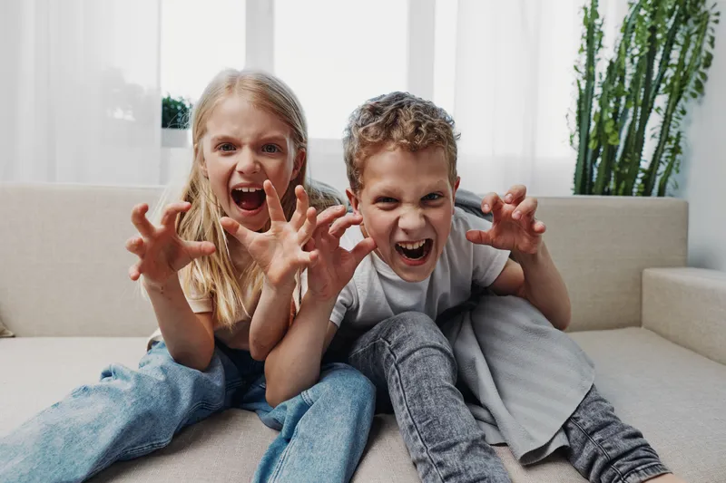 Two playful children sitting on a sofa are making funny claw gestures and faces, showing joyful energy and sibling fun in a bright living room setting.