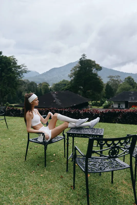 Athletic woman relaxing in white sportswear on a garden chair, surrounded by lush greenery and mountains, conveying a sense of tranquility and fitness
