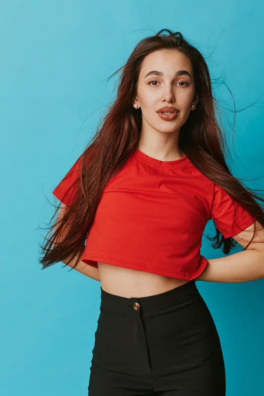 Young woman with long brown hair wearing red cropped t shirt and black pants posing confidently against blue background in studio portrait. Casual style and natural makeup look.