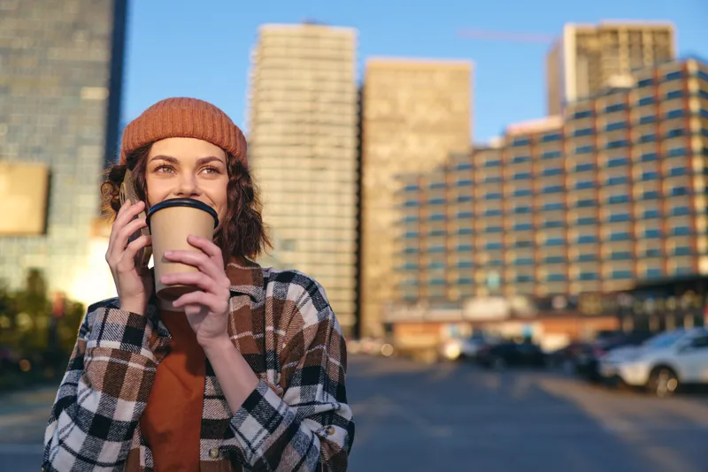Woman with coffee and phone in urban street, candid lifestyle portrait in a beanie and plaid jacket during golden hour glow. Authenticity, mindful living and emotional storytelling in a relaxed city