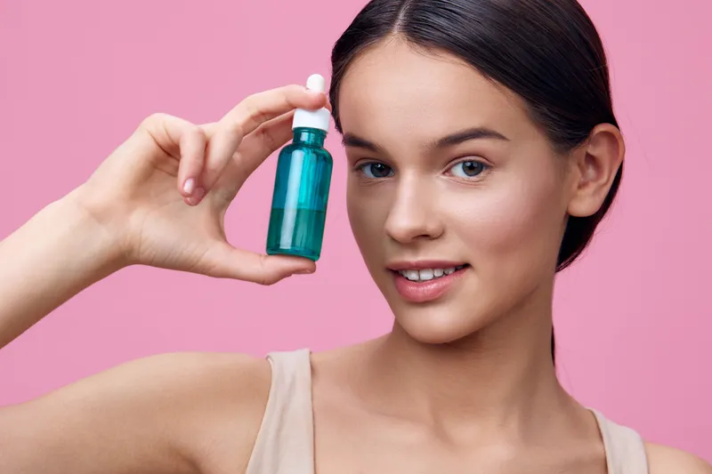 Young woman with smooth skin holding a green serum bottle on a pink background, showcasing skincare routine and healthy beauty concept