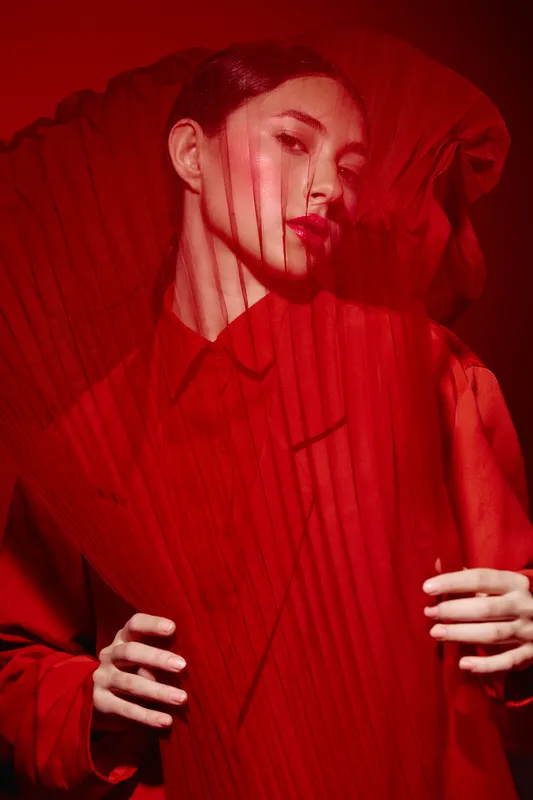Woman in bold red fashion dress and folded fan creates a stylish creative beauty portrait with strong shadows. Artistic studio shot focused on form and texture contrast.