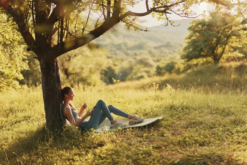Young woman enjoying a peaceful moment under a tree in nature, wearing casual attire, capturing a serene and relaxed atmosphere, with a vibrant background