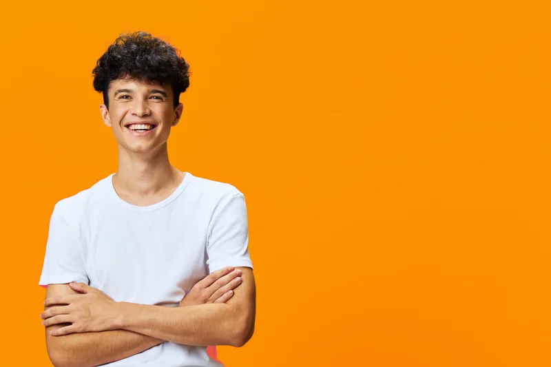 cheerful young man smiling with crossed arms against bright orange background, casual white t-shirt, curly hair, positive energy, studio portrait