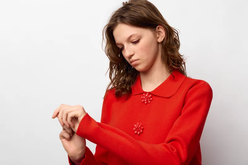 Young woman in red sweater looks down at her wrist with a thoughtful expression. Studio portrait on white background. Concept of contemplation, fashion, and casual style.