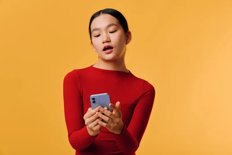 Young woman in red sweater looking at phone with focused expression on yellow background, technology use, communication, digital device, casual style and modern lifestyle concept.