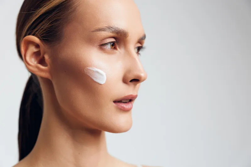 Young woman with clear skin applies moisturizing cream on her cheek while looking thoughtfully to the side against a plain light background.