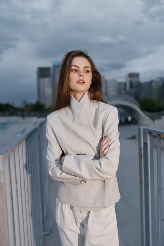 Woman in light gray jacket posing confidently against urban skyscraper background, natural makeup, brown hair, overcast sky, modern fashion style, business theme