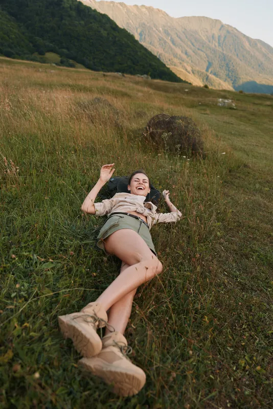 Smiling woman lying in a grassy field with mountains in the distance, enjoying a sunny outdoor moment. Casual, relaxed lifestyle in nature, showcasing freedom, comfort, and a connection to wide