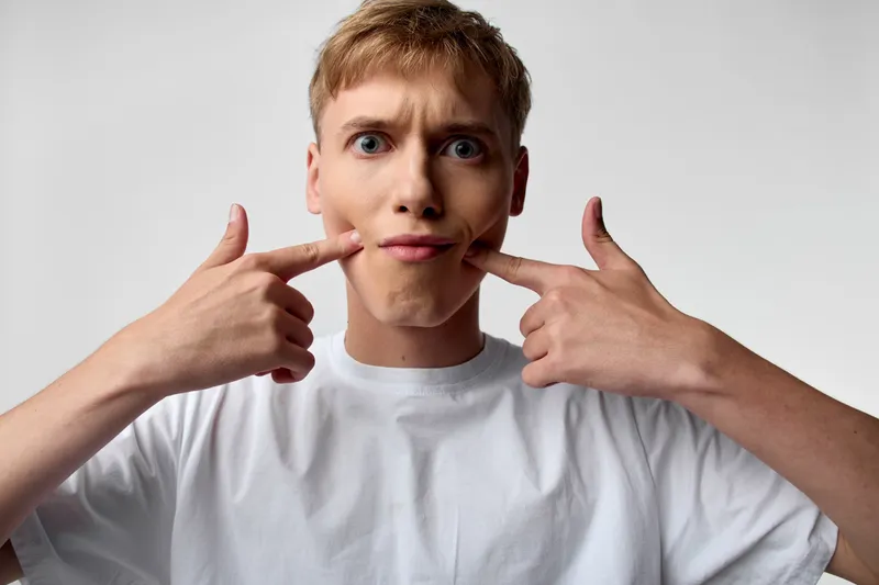 Funny expression, facial distortion, playful pose, mouth corners, cheek stretch, casual shirt, light background, studio shot, entertainment mood, humorous face in a youthful moment, candid vibe and