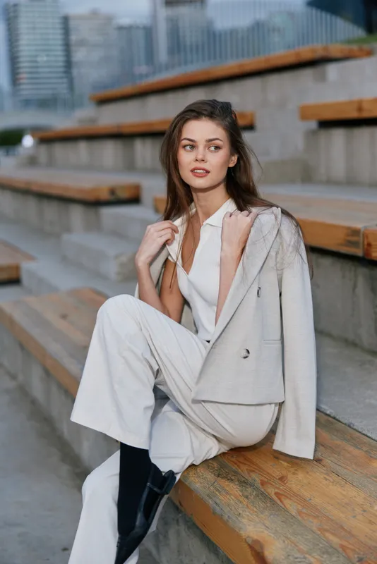Confident businesswoman in stylish outfit sitting on outdoor steps, showcasing modern attire and vibrant city background, exuding professionalism and elegance.