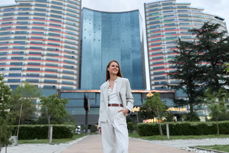 Confident business woman in white suit walking outdoors with modern skyscrapers and greenery background, smiling and relaxed in urban setting