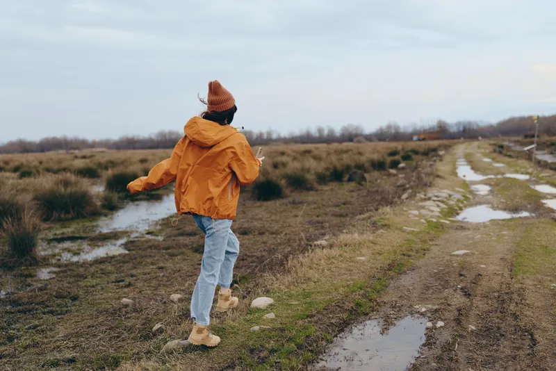 Woman lifestyle outdoors, walking on muddy path through grassy field, orange jacket and knit hat, blue jeans and boots, running shoes, overcast daylight, calm mood countryside with trees
