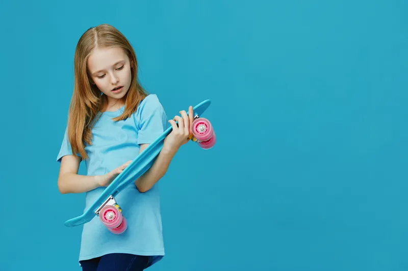 A young girl with long hair wearing a blue shirt examines her turquoise skateboard on a vibrant blue background, embodying joy and curiosity in a playful atmosphere