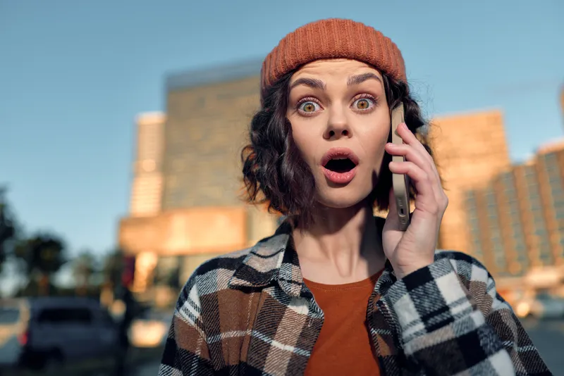 Woman on phone with surprised expression in urban portrait, beanie and plaid jacket outdoors in golden hour glow. Candid lifestyle image conveying authenticity and emotional storytelling.