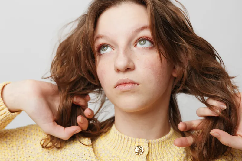 Young woman with curly brown hair wearing yellow sweater looks thoughtfully upward. Studio portrait on white background with natural expression and soft lighting. Concept of contemplation.