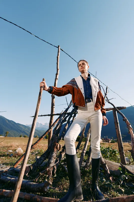 Woman, jacket, boots, stick, mountains, rural — stylish young woman in shearling jacket and white pants holding a stick by rustic wooden structure in mountains meadow landscape.