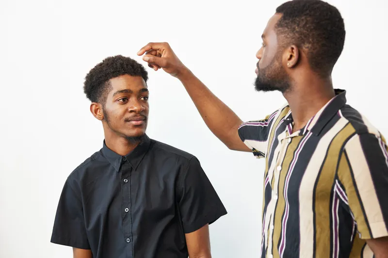Two black men share a moment of camaraderie, showcasing friendship and style against a clean, minimalist backdrop. Their expressions reflect warmth and connection.