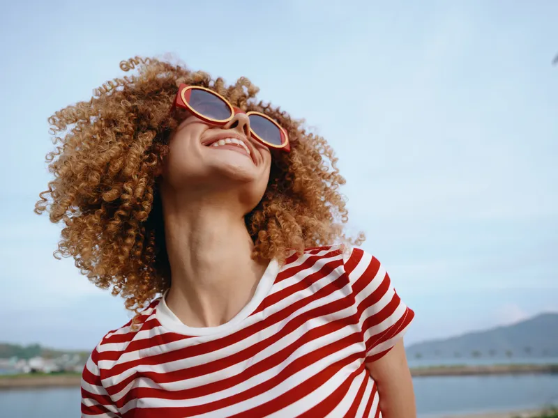 Joyful portrait of a young person with curly hair wearing a red striped shirt and round sunglasses, basking in bright sunlight by a calm seaside backdrop.