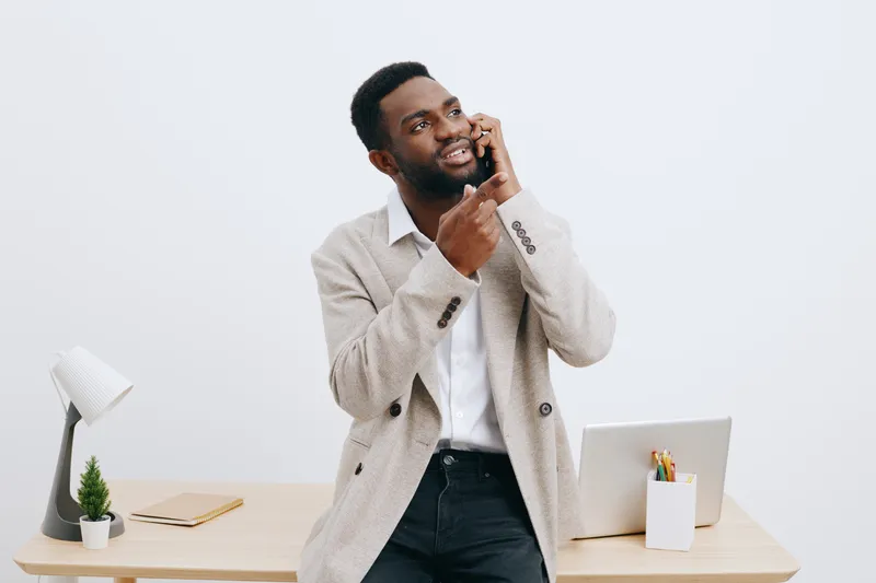 Friendly smiling man with beard in casual beige blazer and white shirt stands at desk with white background, talking on mobile phone, confident and relaxed, office workspace. People lifestyle concept