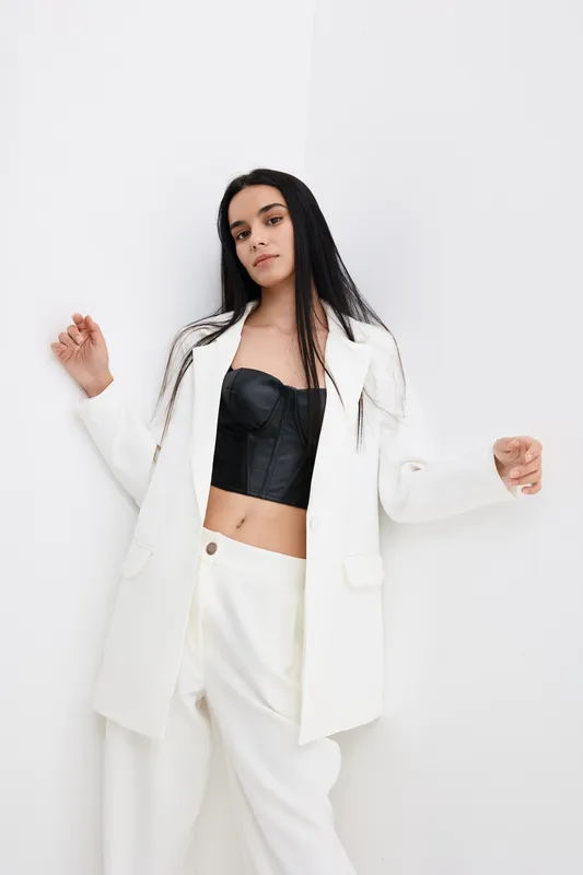 Young woman in white blazer and trousers posing confidently against plain white wall, wearing black corset top with long straight hair, minimalistic fashion style.