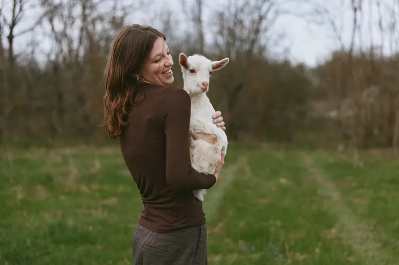 Young woman in brown sweater happily holds a white baby goat outdoors in a green field during early spring, enjoying a peaceful moment with the animal in natural light.