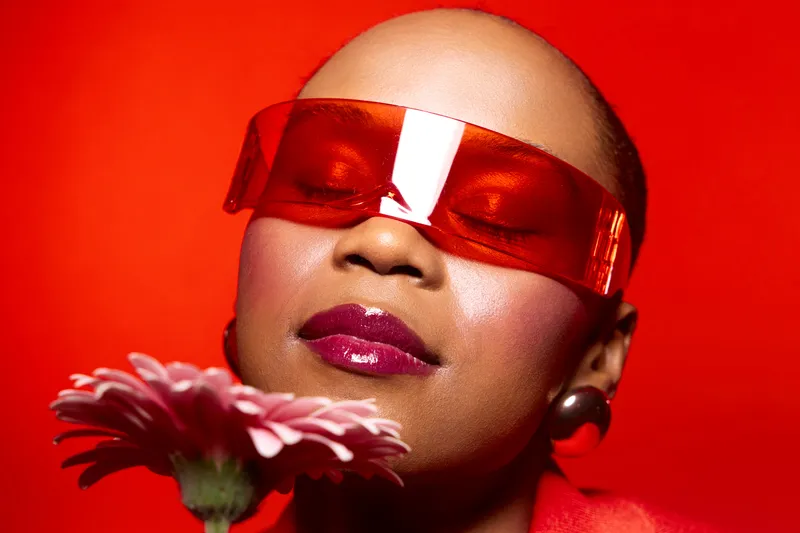 Close-up of a dark skinned African woman with bald head wearing red futuristic sunglasses holding pink flower. Concept of bold fashion, beauty, and modern style on red background.