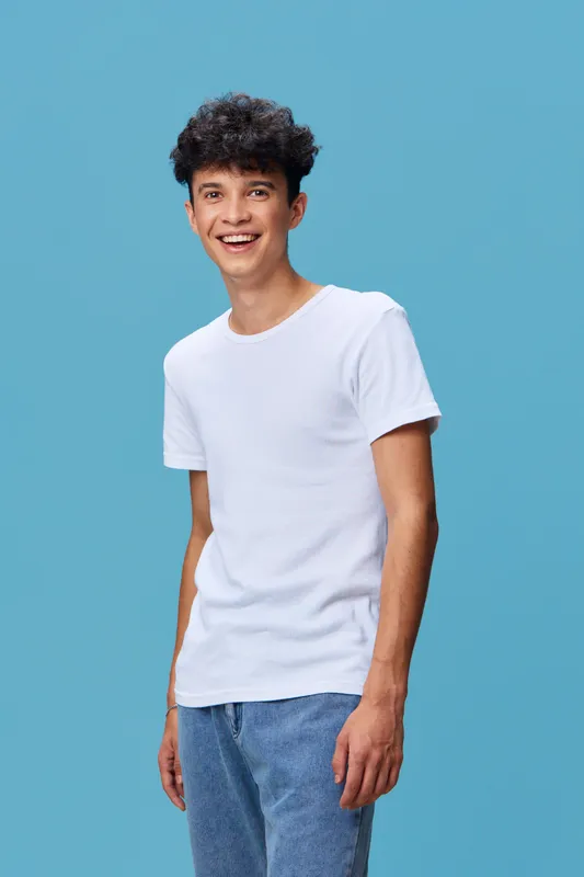 smiling man wearing casual white t-shirt and denim jeans stands against blue background, expressing happiness and youthfulness in a studio setting