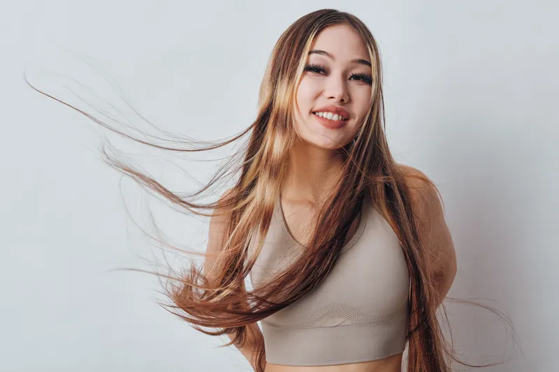Young woman with long flowing hair smiling and posing in sleeveless top on plain light background. Female model happy and confident with natural look and casual style.