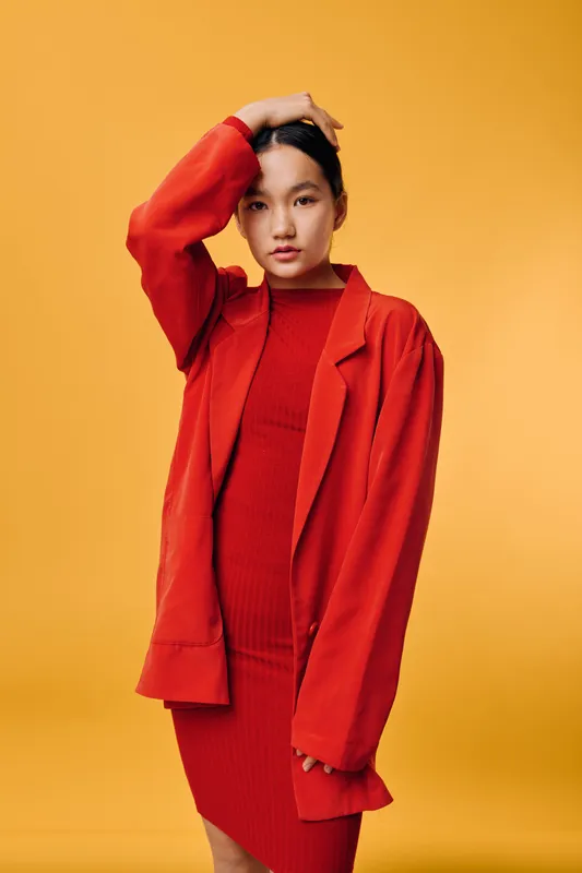 Young woman posing in red dress and oversized jacket on yellow background, fashion studio portrait with confident gaze and modern casual style in minimal setup lighting.