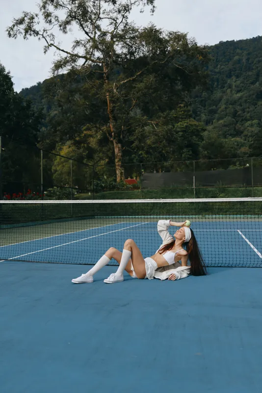 Young female tennis player relaxing on the court, showcasing athletic wear, emotion, and vibrant outdoor scenery, surrounded by green trees and natural light