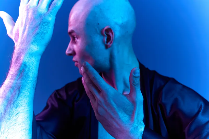 A striking portrait of a man with a bald head in a vibrant blue light, showcasing an intriguing expression and artistic pose, conveying emotion and depth.