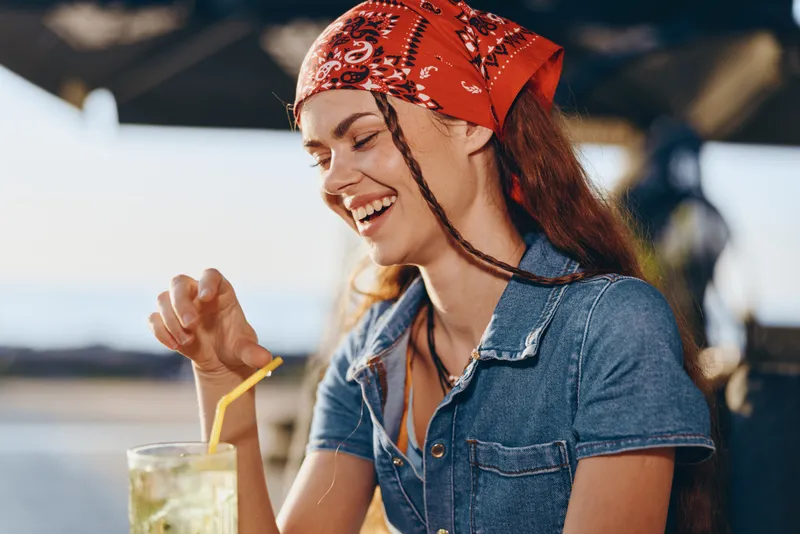 Lifestyle woman in modern boho-western denim street style enjoys refreshing drink outdoors with film color tones enhancing warm, casual, and joyful atmosphere in sunny setting.