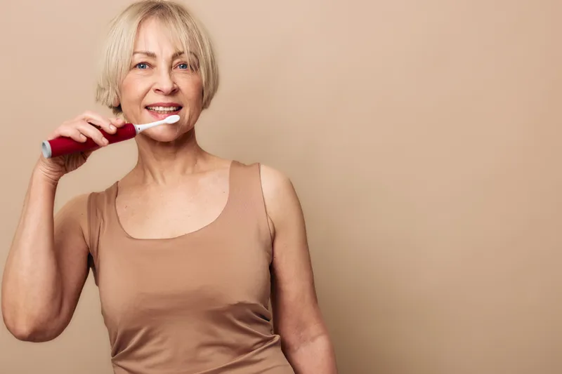 Mature woman brushing teeth with electric toothbrush against beige background. Healthy dental care, personal hygiene routine, oral health concept for senior woman in casual wear.