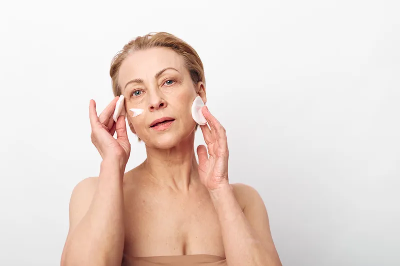 Mature woman applies face cream and uses cotton pads for skincare on clean skin. Close-up with neutral background shows beauty, self-care, anti-aging, and natural skincare concept.