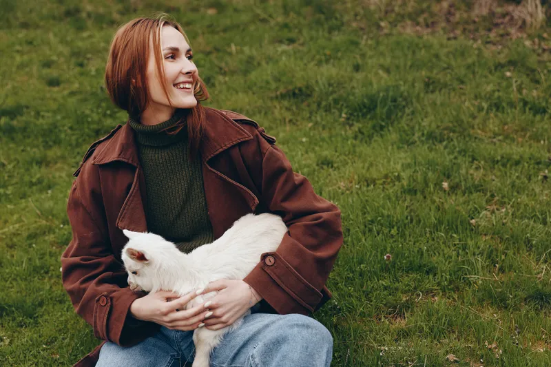 Cheerful woman outdoors cuddling a white goat kid, smiling softly, sunlit meadow scene, relaxed pose, natural colors, rustic outfit, peaceful countryside moment