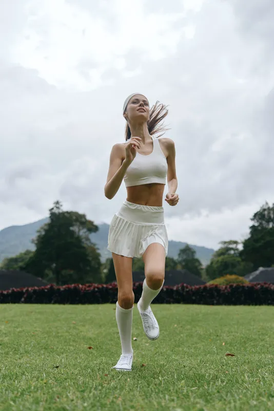 Young athletic model running on a green field in sporty white clothing, showcasing energy and freedom with a cloudy sky background