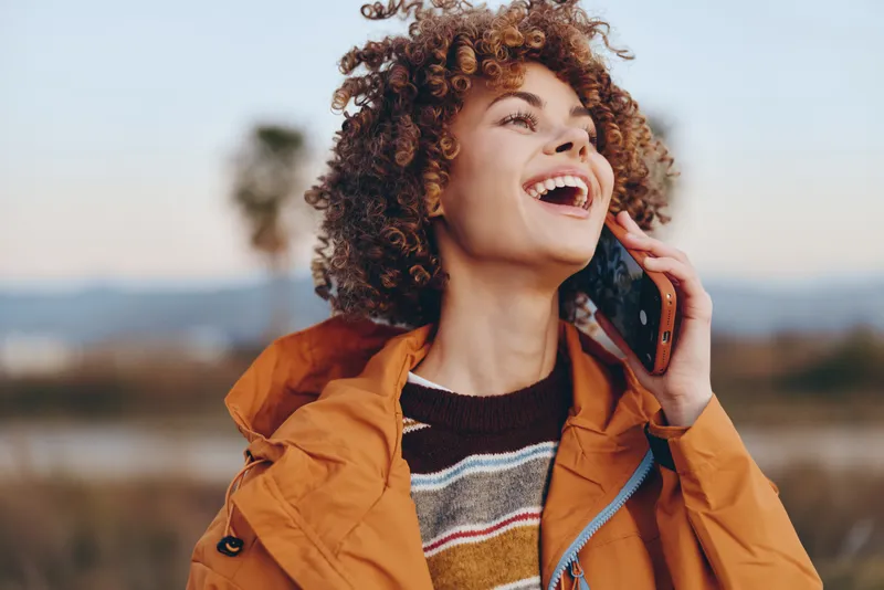 Lifestyle woman outdoor autumn landscape, curly hair and orange jacket, smiling and speaking on phone to ear, making call with warm sunlight, casual travel mood in fresh air today.