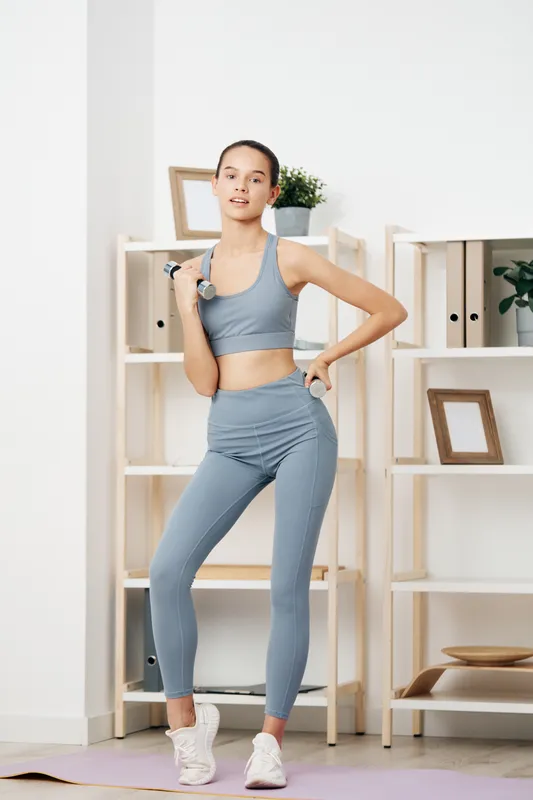 Woman in athletic wear performing a dumbbell curl in a bright home gym, showcasing fitness, strength training, balance and focus for a healthy lifestyle. She stands on a mat beside shelves with