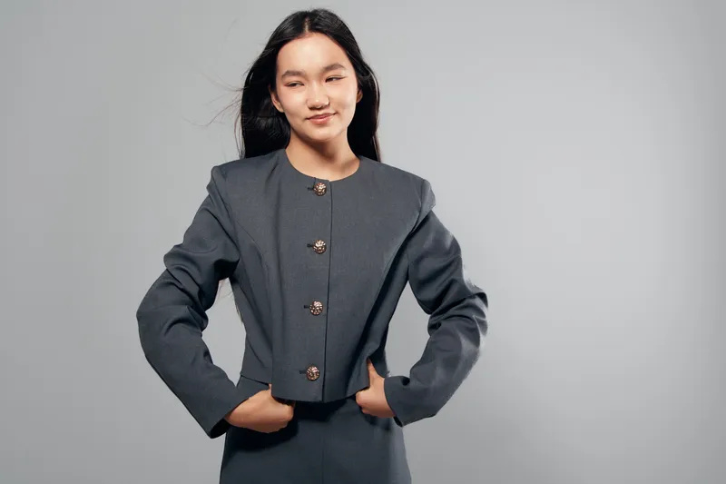Young Asian woman in gray formal suit with buttons posing confidently with hands in pockets against plain background. Female model with long dark hair and slight smile in studio.