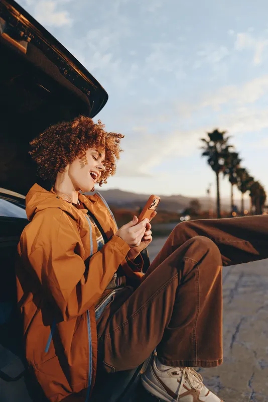 Woman lifestyle scene outdoors near a parked car, curly haired woman relaxes on the road, using a smartphone and smiling in warm daylight, travel mood, casual fashion