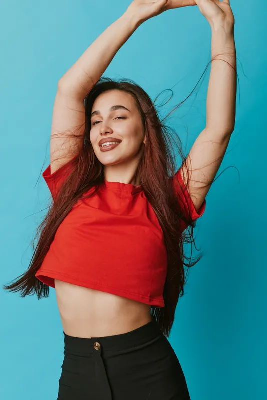 Young woman in red crop top and black pants posing with arms raised, smiling confidently with closed eyes against blue background. Fashion, beauty and lifestyle portrait.